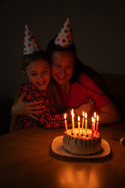 一位戴着派对帽的母亲拥抱女儿，旁边是点亮的生日蛋糕，在昏暗的家中温馨的夜晚庆祝活动中，分享着喜悦和亲情的温暖节日时刻。竖构图照片图片下载