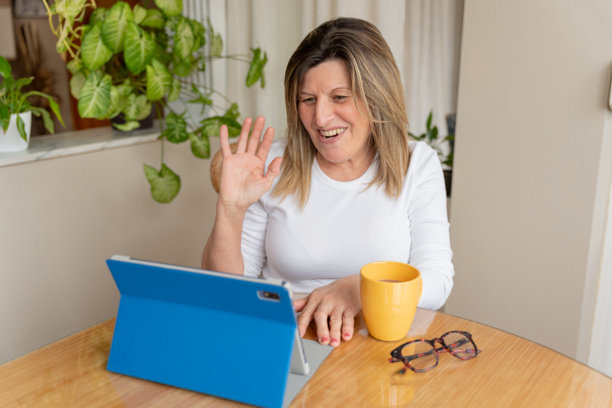 一位成熟的成年女性在视频会议中对着平板电脑屏幕微笑挥手，享受远程沟通的乐趣图片下载