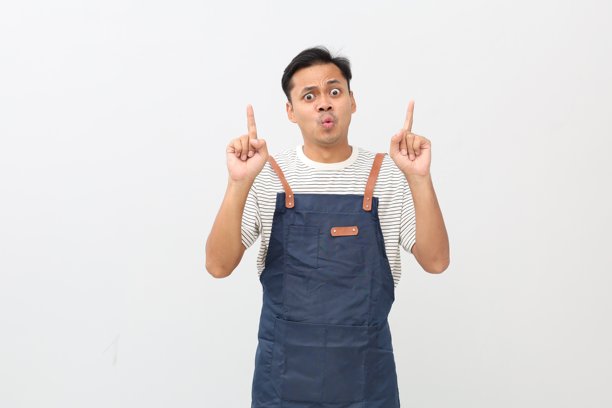 惊讶的亚洲男性的服务员穿着蓝色围裙，展示着标志或广告，向上竖起手指指向复制空间，站在白色背景上图片下载