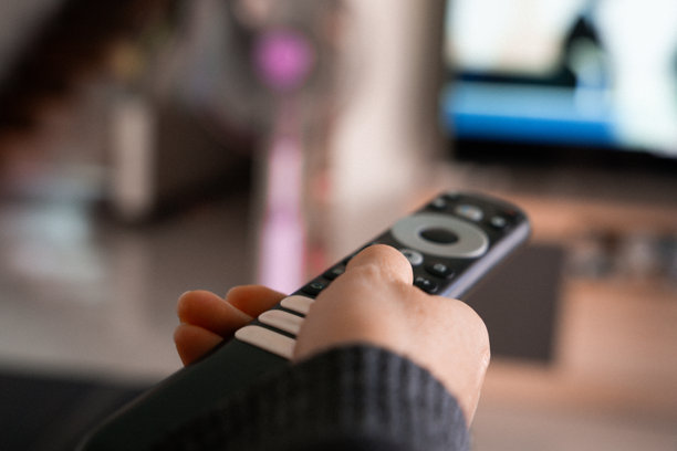 特写一只手拿着电视遥控器，背景是模糊的客厅和电视屏幕。体现家庭娱乐、科技使用和现代生活方式的概念。图片下载