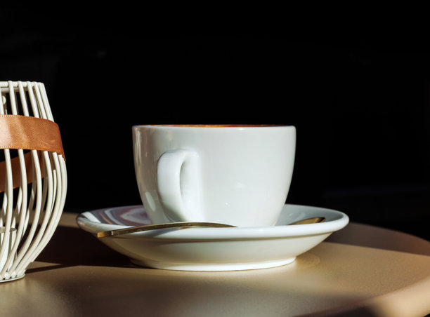 阳光下的咖啡馆桌子，一个白色的咖啡杯放在碟子上，旁边放着勺子，背景为黑色，带有大片黑色留白空间图片下载