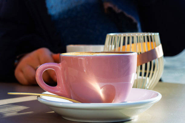 阳光明媚的日子里，咖啡馆的桌子上放着两杯咖啡，其中一杯卡布奇诺上面有心形拉花，背景中坐着一个人。焦点在前景。图片下载