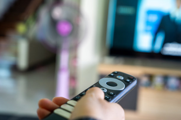 特写一只手拿着电视遥控器，背景是模糊的客厅和电视屏幕。代表家庭娱乐、技术使用和现代生活方式的概念。图片下载