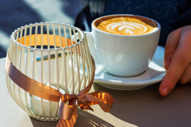 阳光明媚的咖啡馆场景，旁边放着一个装饰有丝带的烛台，一杯卡布奇诺咖啡上有着拉花。背景中有一只人的手，焦点经过选择。图片下载