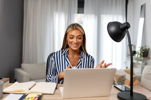 年轻的女商人在家庭办公室的笔记本电脑上进行视频通话。微笑的女士在在线会议或网络研讨会中做出手势并交谈。远程沟通、虚拟会议和数字工作空间。图片下载