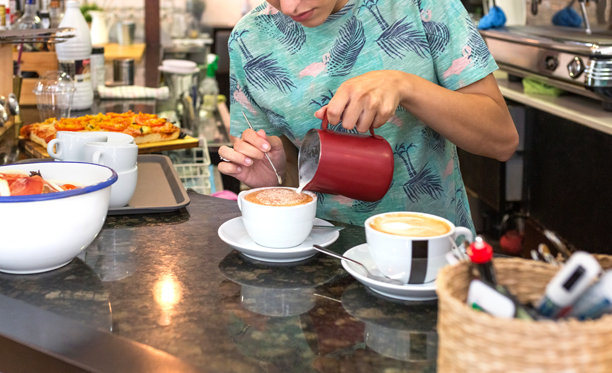 年轻的女咖啡师从奶罐中倒出打发好的牛奶，在卡布奇诺上制作拉花图片下载