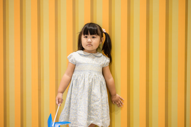 可爱的亚洲小女孩，手持蓝色风车，站在复古黄条纹墙前，画面捕捉了一位身穿蓝色碎花裙的小女孩，手持蓝色纸风车，对着镜头露出迷人笑容，背景是略显陈旧的黄橙相间条纹墙，整体散发出童趣与怀旧的气息。图片下载
