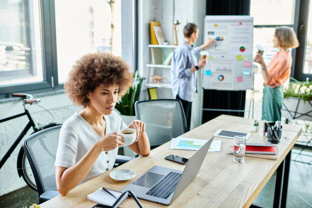 女性在现代办公室空间协作，其中一人享用咖啡并专注于笔记本电脑图片下载