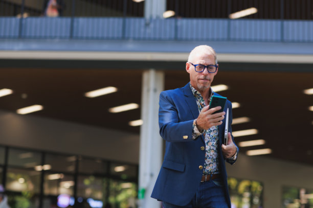 成熟男子一手拿着笔记本电脑，一手查看智能手机，行走在城市街道上图片下载