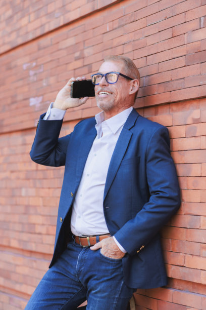一位成熟的男士带着微笑，站在砖墙前，正在用智能手机沟通。图片下载