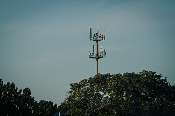 在晴朗蓝天下，一座拥有天线的通信塔高耸于郁郁葱葱的绿树之上，展示了城市环境中现代化的电信基础设施，并留有复制空间图片下载