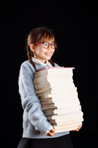 小女孩在黑色背景的影棚里，怀里抱着一大摞书，摆拍照片。图片下载