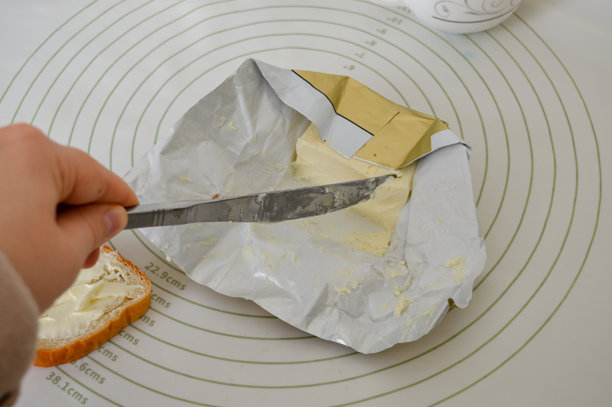 特写照片，一只手正用刀在新鲜的面包片上涂抹黄油。简单的自制早餐场景，自然光，日常食物准备概念。图片下载