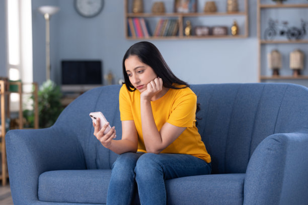 漂亮的年轻女孩穿着休闲服装，在家坐在沙发上玩手机的素材照片图片下载