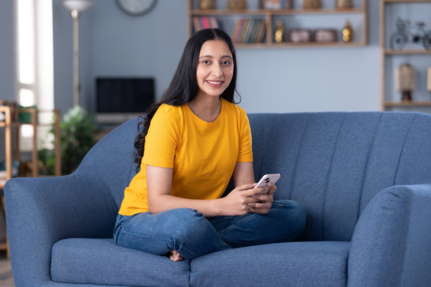 身穿休闲服饰的漂亮年轻女孩在家中沙发上使用手机的库存照片图片下载