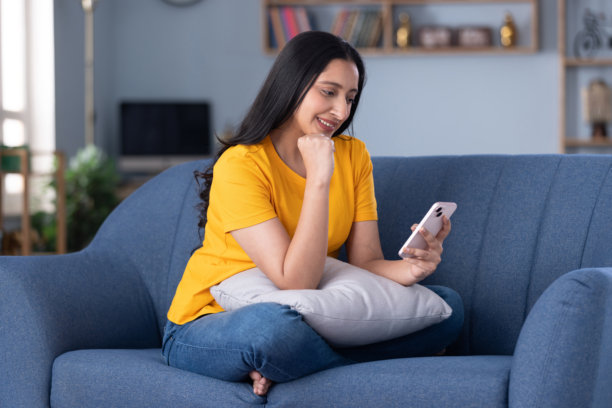 穿着休闲服饰的漂亮年轻女孩在家中沙发上使用手机的居家照片图片下载