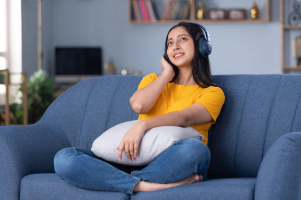 照片：一个漂亮的年轻女孩在家中沙发上，戴着耳机，微笑着享受音乐图片下载