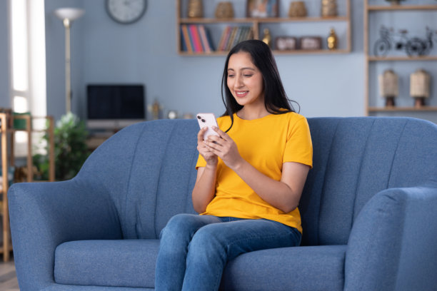 穿着休闲服的年轻美女坐在家里的沙发上玩手机的真实照片图片下载