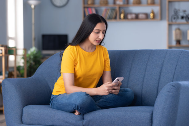 身穿休闲服饰的漂亮年轻女孩在家中沙发上使用手机的居家照片图片下载