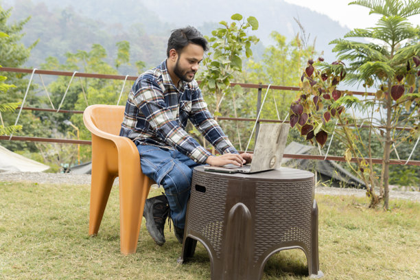 印度年轻男子在花园里远程办公，坐在户外椅子上，在度假时使用笔记本电脑。图片下载