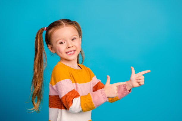 一位年轻女孩穿着色彩条纹毛衣，对着明亮的蓝色背景露出微笑，并用双手指向前方，充满活力的家庭生活与时尚素材照片图片下载