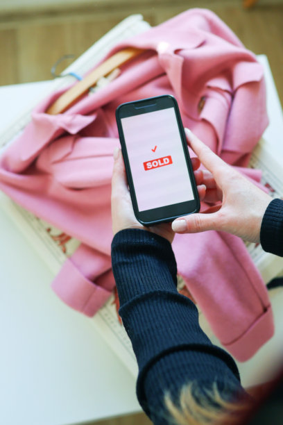 粉色衣服上的智能手机屏幕显示已售出确认。网购、销售成功和二手时尚概念。图片下载