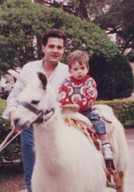 1980年代照片：一位年轻的父亲带着儿子，父亲骑在羊驼上摆姿势图片下载