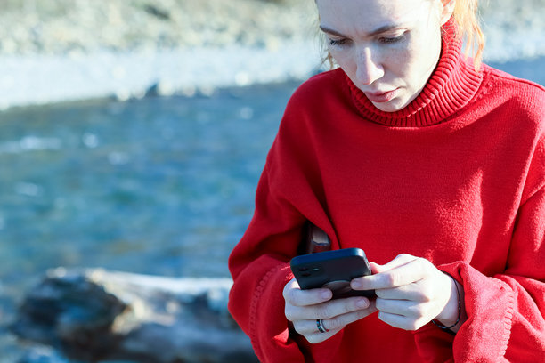 身穿红色高领毛衣的女子在河边使用智能手机，专注于屏幕，背景是自然风光。图片下载