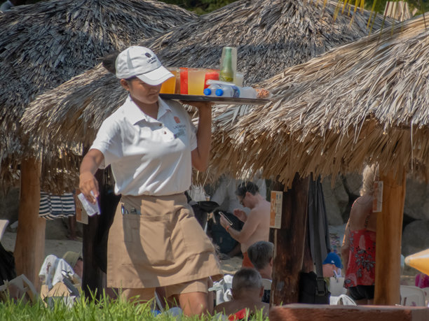 一名服务员在阳光明媚的海滩俱乐部为客人端着沉重的点心托盘。真实的镜头展示了夏日的待客之道和度假村的服务。图片下载