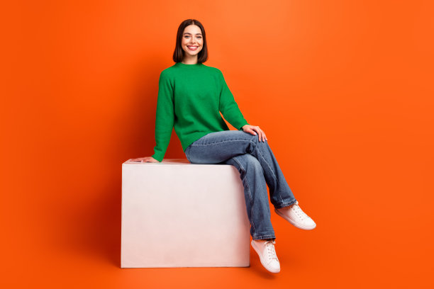 年轻时尚的女性身穿绿色毛衣，坐在白色箱子上，背景为橙色，用于时尚生活方式图库照片图片下载