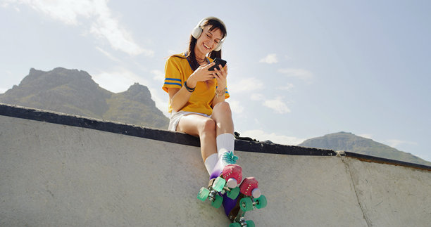女人在公园里，脚穿轮滑鞋，手拿手机，脸上带着微笑，戴着耳机听音乐，背景是城市空间。这个人是轮滑爱好者，用智能手机观看流媒体订阅内容，低角度拍摄，在城市里的斜坡上放松。图片下载