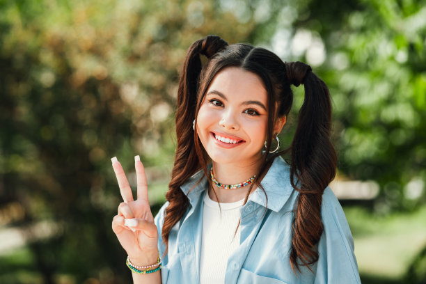 一位年轻女子穿着浅蓝色衬衫，在阳光明媚的公园里做出胜利手势并微笑着，这是一张生活时尚风格的素材图图片下载