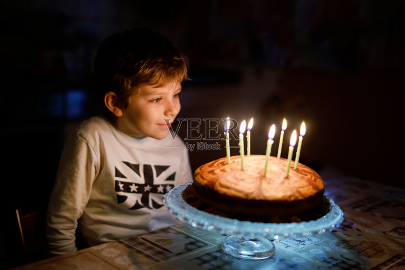 可爱的快乐的金发小男孩庆祝他的生日。孩子吹七根蜡烛在自制的烤蛋糕上，室内。正版图片素材