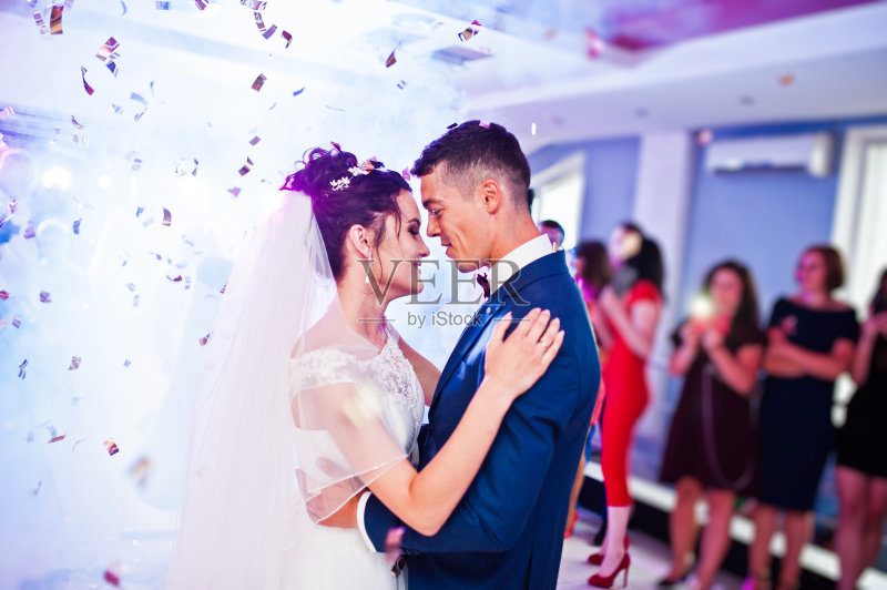 这是新人在婚礼上以五彩纸屑和彩灯为背景的感人的第一支舞。正版图片素材