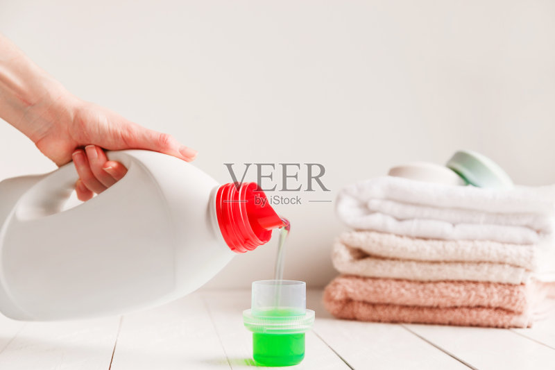 近距离观察女性的双手将洗衣液倒入白色质朴的桌子上，背景是浴室里的毛巾。正版图片素材