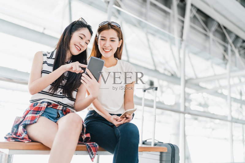 两个快乐的亚洲女孩带着行李一起用智能手机检查航班或在线登机。航空旅行，暑假，或手机应用技术概念正版图片素材