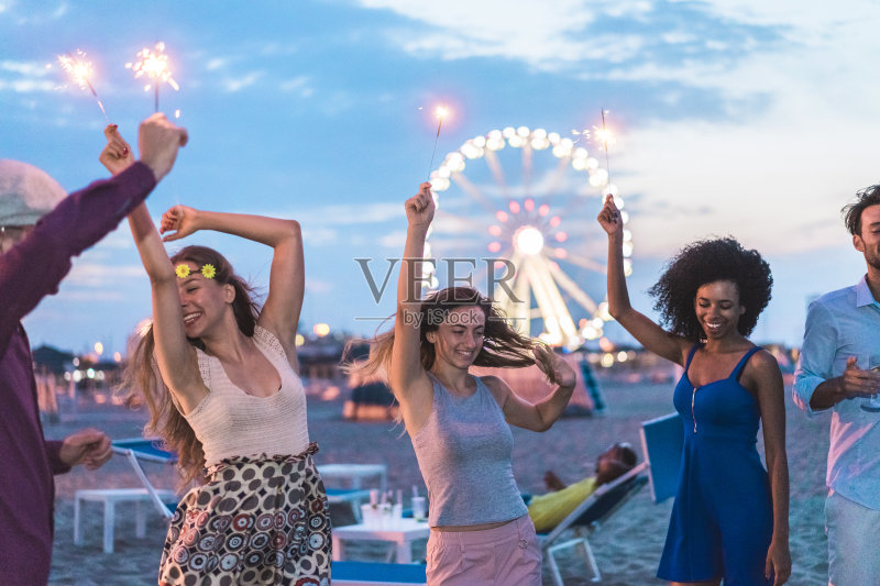 朋友们在日落后举办海滩派对，燃放烟花，喝香槟 - 年轻人在户外跳舞 - 柔和的焦点集中在中间女孩的脸上 - 青春和节日的概念正版图片素材