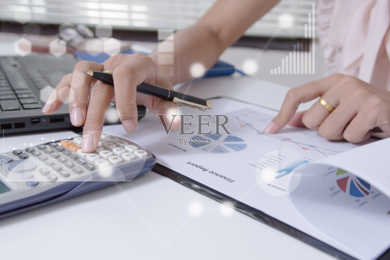 年轻的金融市场分析师坐在白色的桌子前，用笔记本电脑在办公室工作。商人分析文件和手中的计算器。笔记本屏幕上的图形和图表正版图片素材