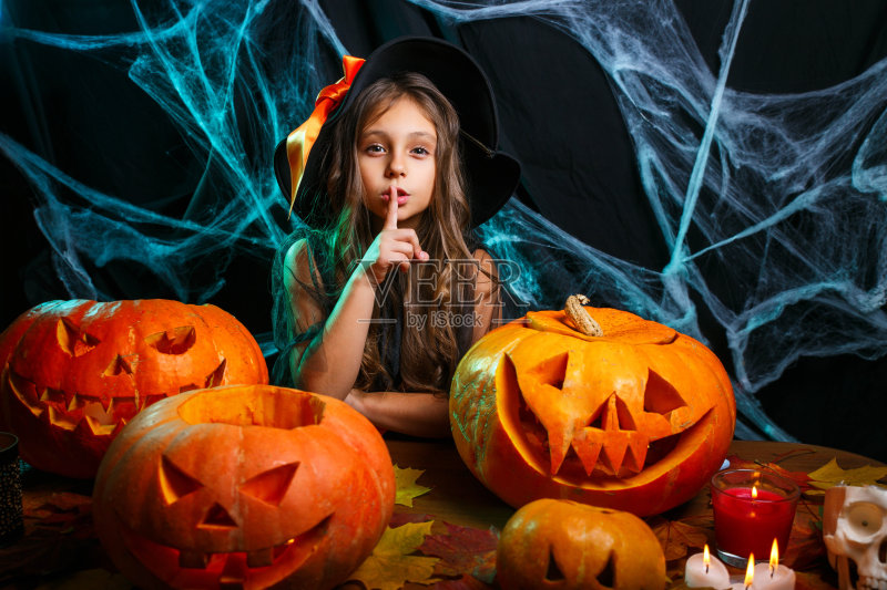 万圣节快乐。小女孩穿着女巫服装庆祝万圣节，用弯曲的南瓜在蝙蝠和蜘蛛网上摆姿势。正版图片素材