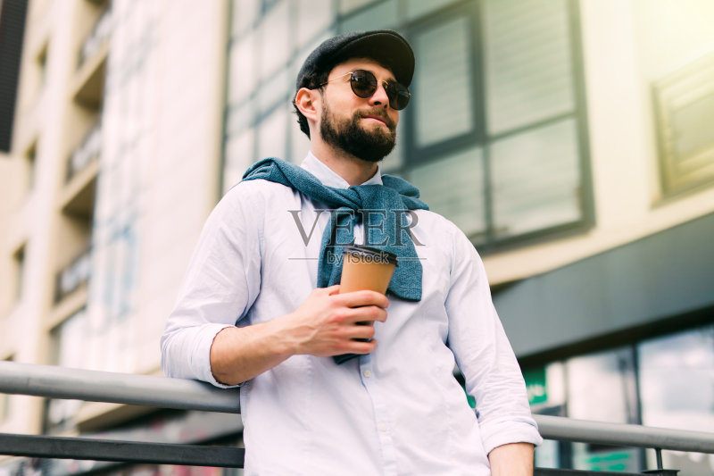 享受新鲜的咖啡。帅气的年轻人穿着休闲服，手拿一次性杯子，微笑着走在城市的街道上正版图片素材