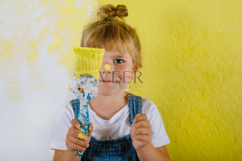可爱的小女孩把新房子的墙刷成了黄色。家庭装修公寓。快乐的小女孩在室内粉刷墙壁，用调色板选择颜色，用画笔玩耍正版图片素材