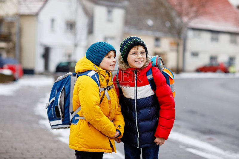 两个小学生男孩在大雪中步行上学。孩子们玩初雪玩得很开心。兄弟姐妹和最好的朋友背着背包，穿着五颜六色的冬装。正版图片素材